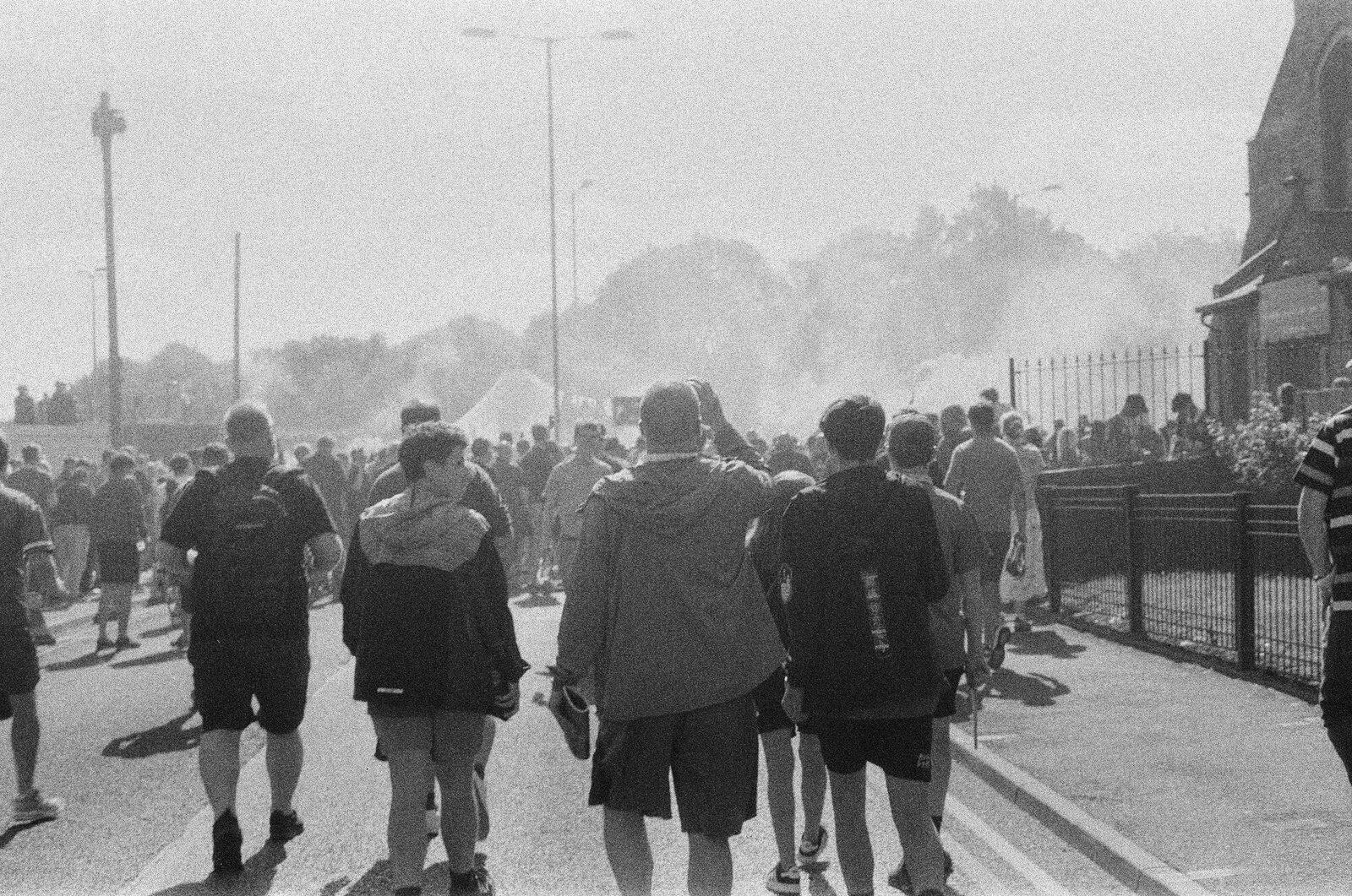 Crowds of fans walking up a sunlit street toward Goodison Park through drifting flare smoke