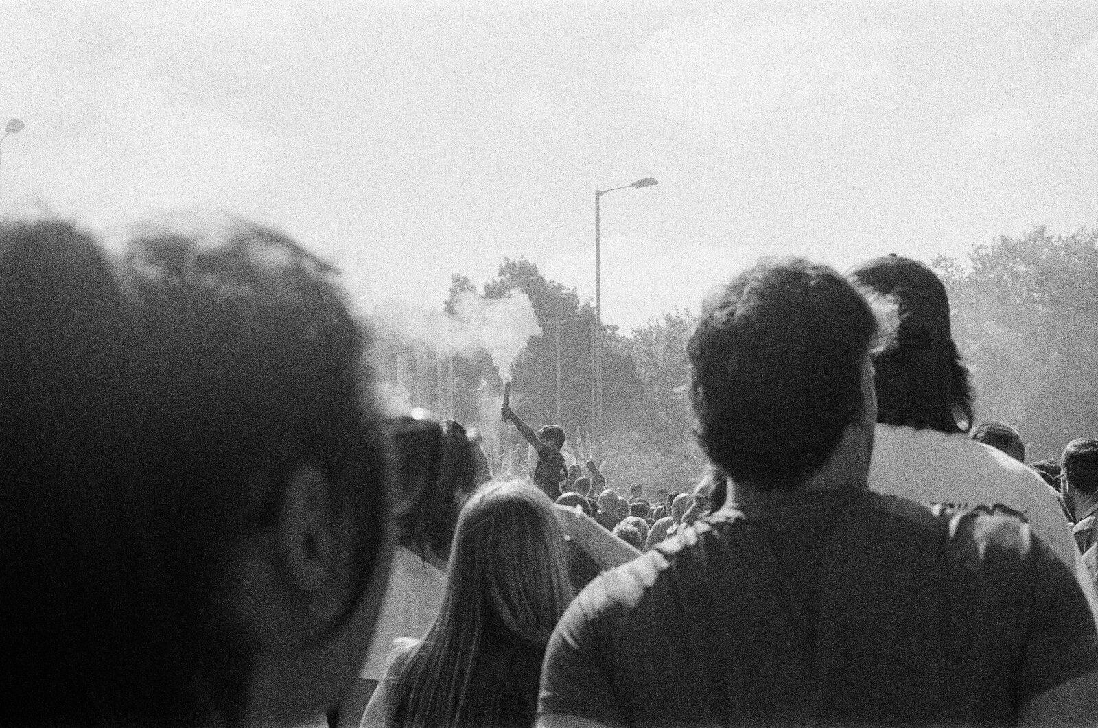 A boy holding a smoking flare up to the sky in the distance, seen between the heads of fans in the crowd