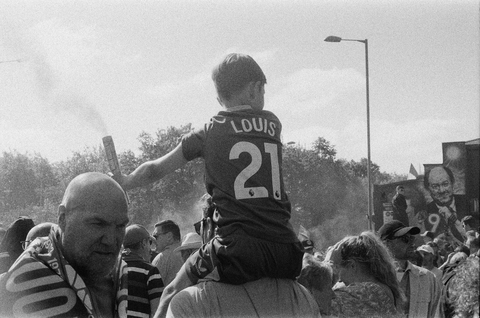 A boy on someone's shoulders in a Louis 21 shirt holding a flare, a banner of a man's face behind