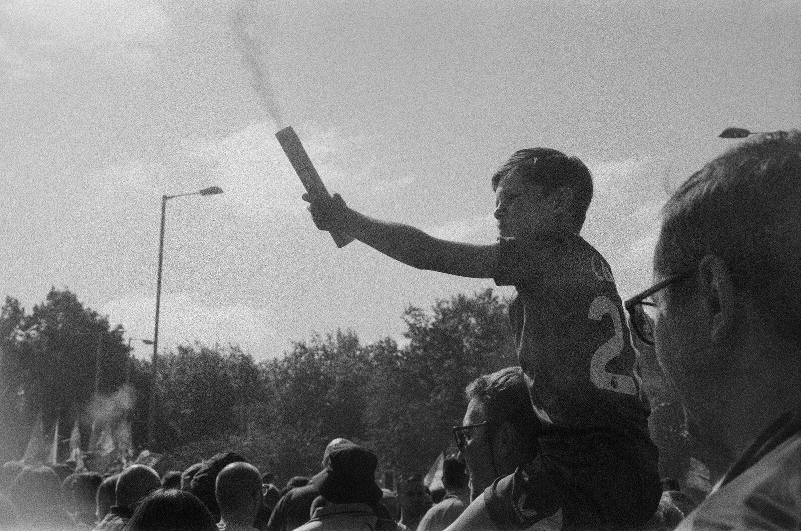 A boy on someone's shoulders holding a smoking flare out at arm's length