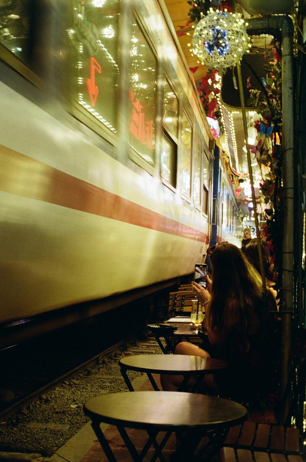 A woman photographing a train passing close to cafe tables at night